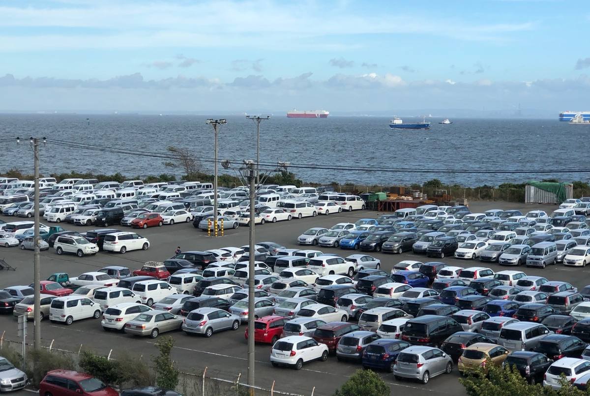 Hundreds of used Japanese cars — kei cars, minivans and hatchbacks — parked row on row in a coastal yard in Japan, with car carrier ships out on the bay behind them