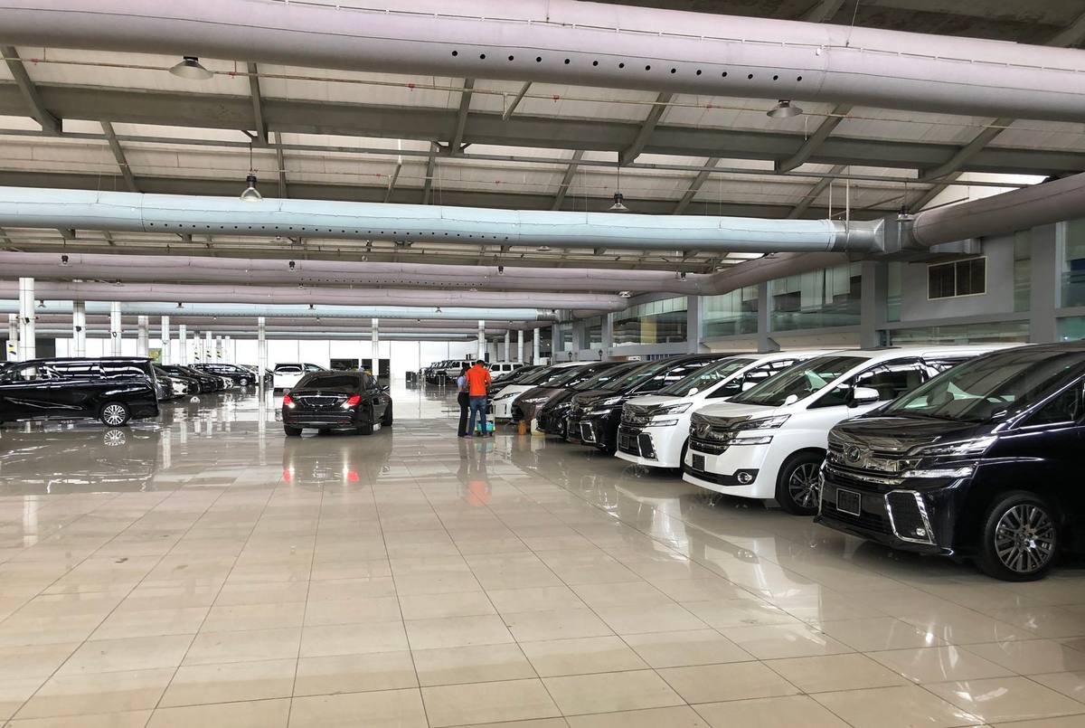 Toyota Alphard and Vellfire minivans and a black Mercedes-Benz sedan lined up on the polished floor of a large indoor showroom, with two people looking over a car in the middle of the hall