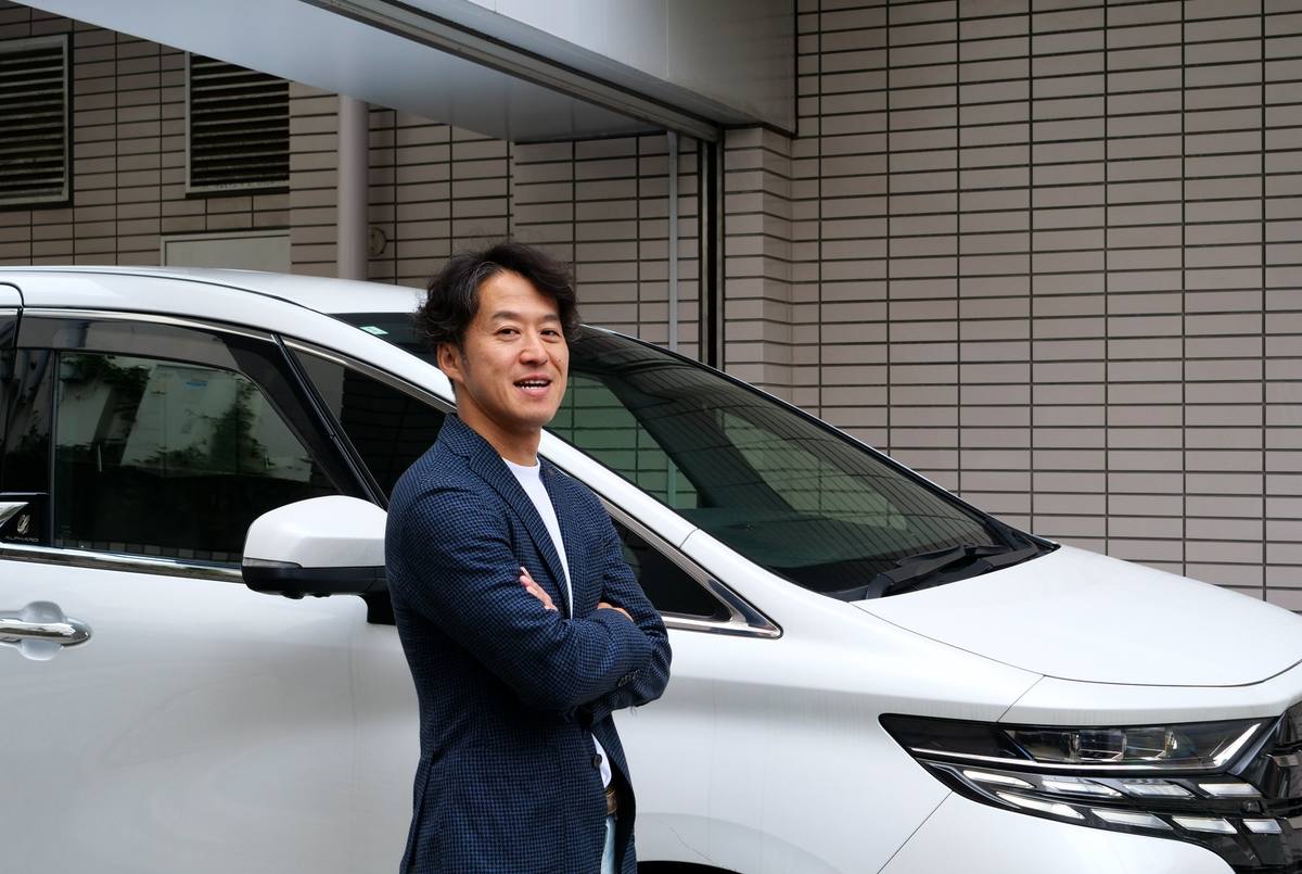 Nobuaki Chatani, Representative Director of UCWORLD, standing with his arms crossed in front of a white Toyota Alphard