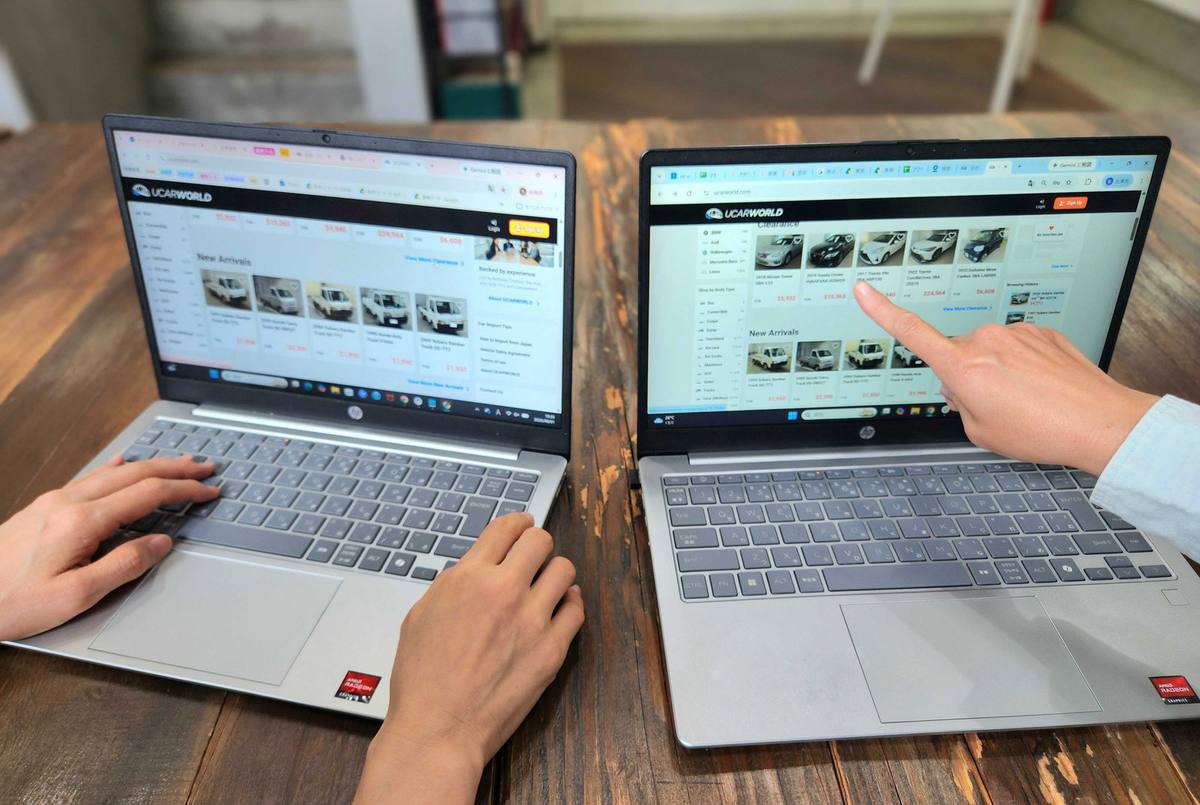 Two laptops on a wooden desk showing the UCARWORLD car listings, with a team member pointing at a vehicle on screen