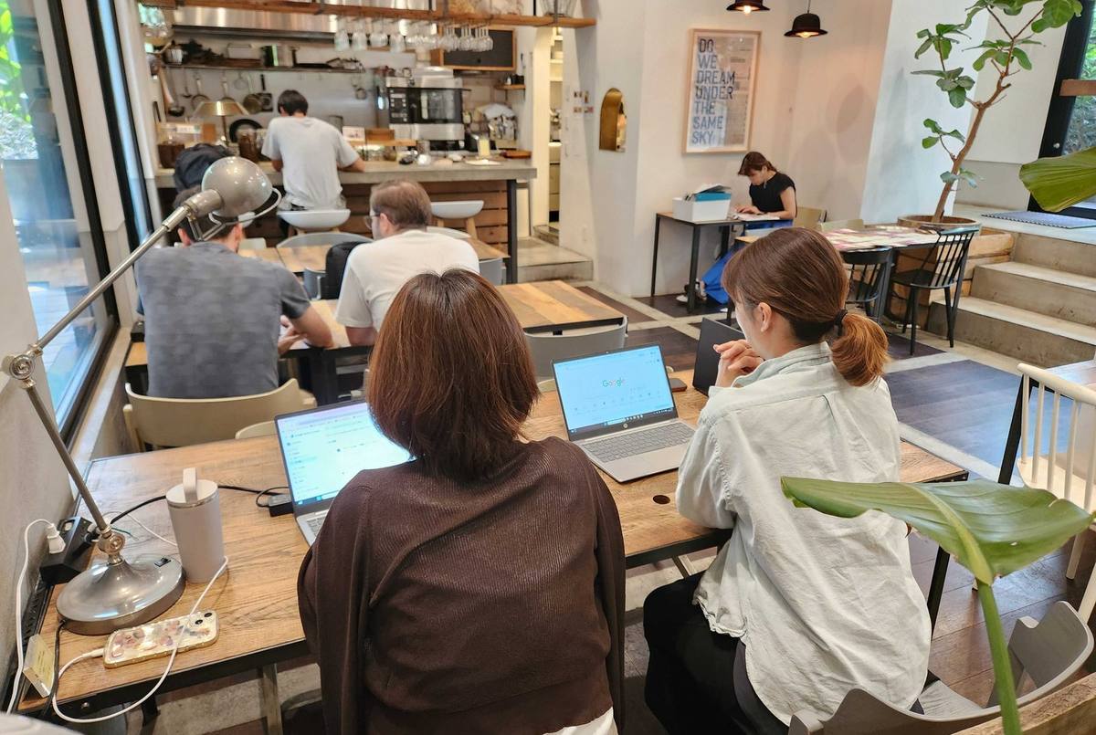Two UCARWORLD staff members working side by side on laptops at a shared wooden table in a bright cafe workspace, with other people working at tables behind them