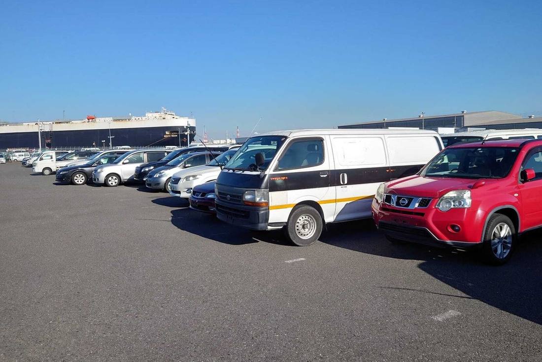Japanese used cars, including a Toyota HiAce van and a red Nissan X-Trail, lined up at a port yard in Japan with a car carrier ship moored behind them